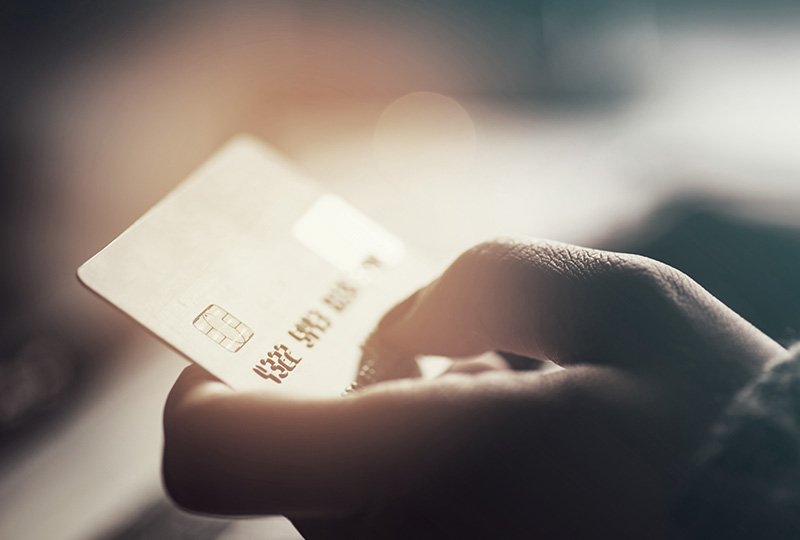 The image shows a close-up of a hand holding a credit card with the focus on the card s details, set against a blurred background that suggests an indoor setting.